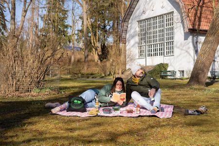 2 Menschen picknicken auf einer Wiese vor einem Haus